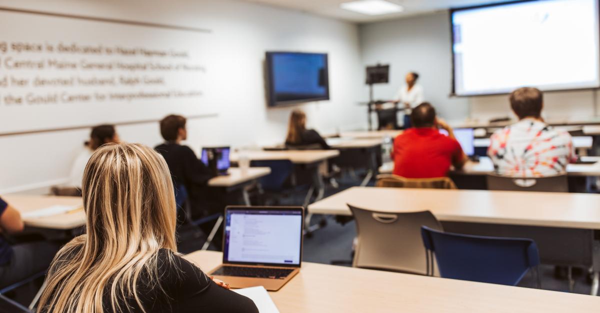 College students seated in a classroom looking at a projector screen while a professor lectures.