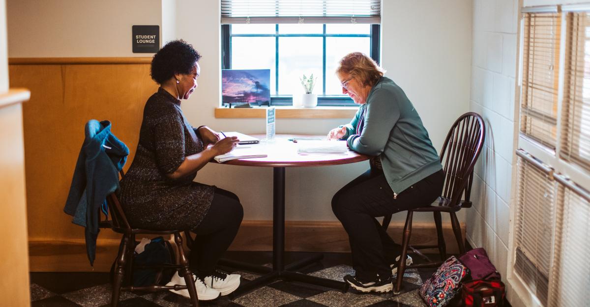 Two students sit at a small table together, studying.  Behind them is a sign that says, 'student lounge.'